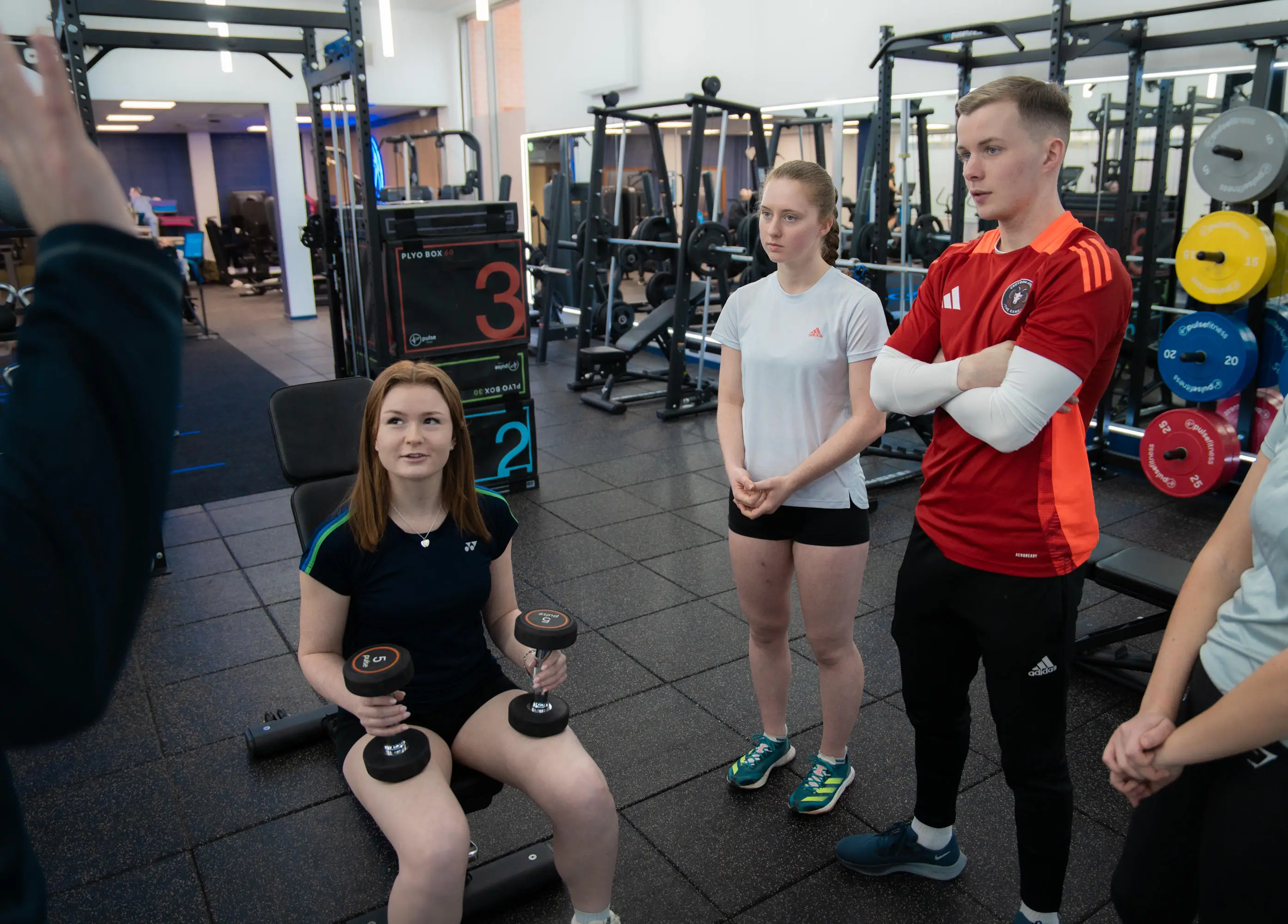 A participant on a bench with weights looking to the trainer with group of people around her
