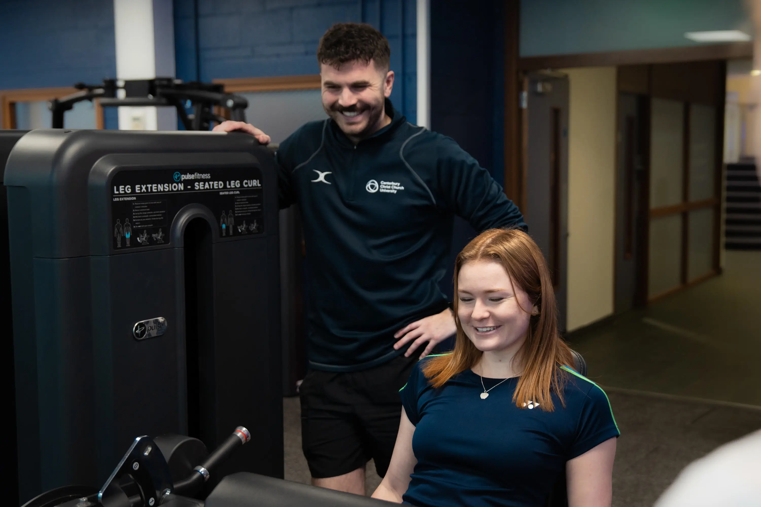Client using machine with trainer stood behind her watching