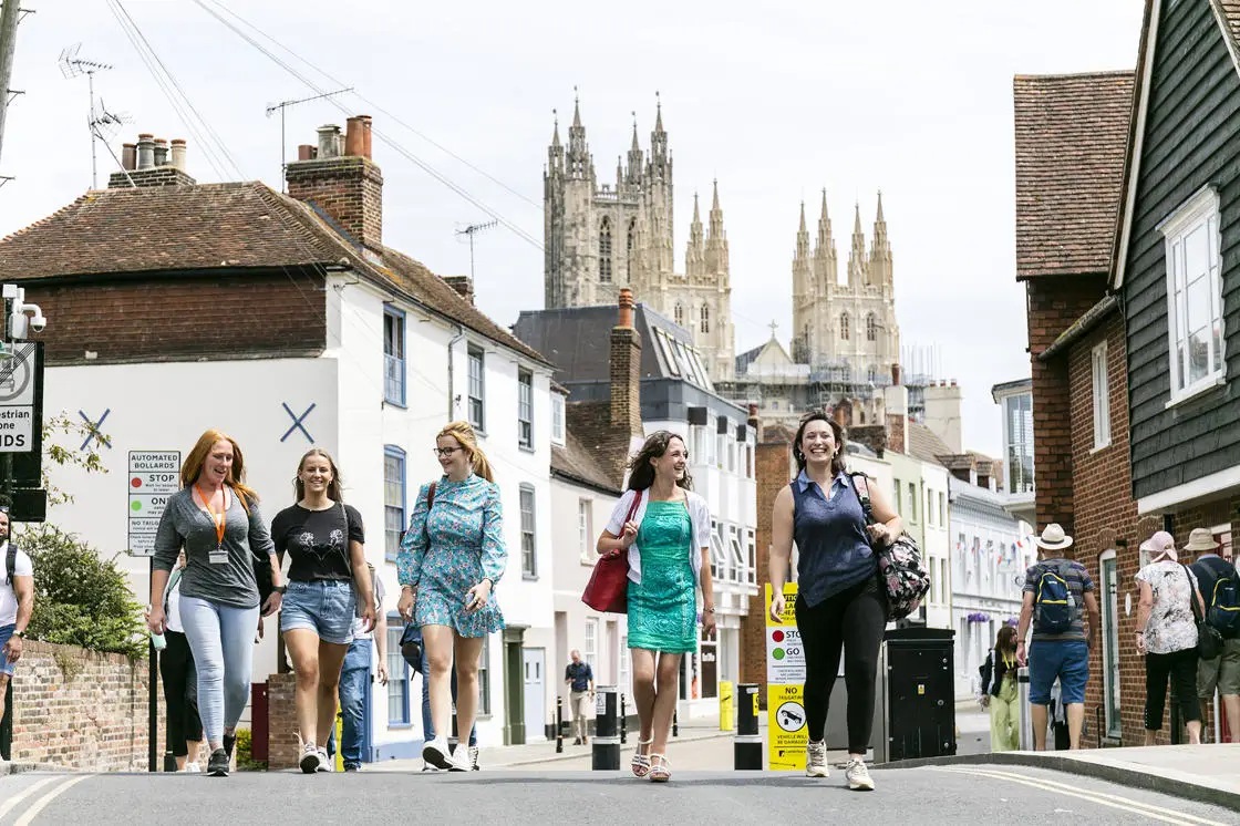 Canterbury Cathedral is visible in the background as a group of visitors walk through the ancient city. 